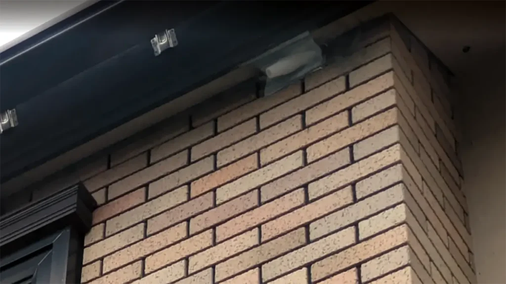 レンガ調の建物外壁と屋根の接合部にある隙間に取り付けられた、コウモリ追い出し用の一方通行デバイス「バットアウト」の設置画像。