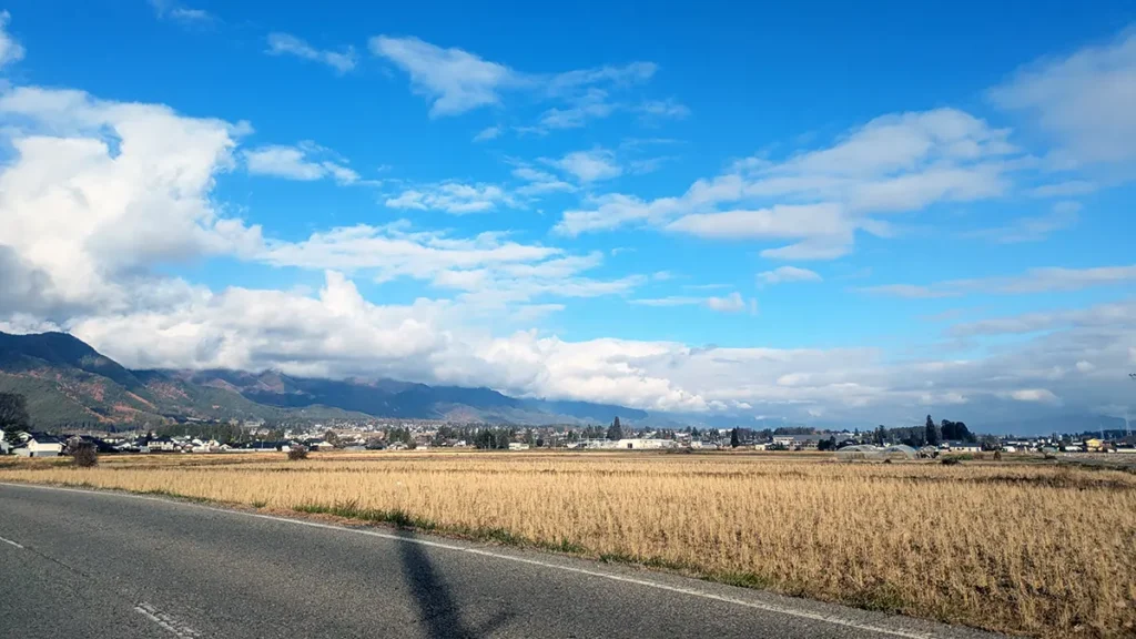 長野県の農村風景。手前に道路と収穫後の田んぼ、奥に住宅地と山並みが広がる。